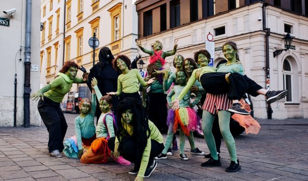 fot. J. Smoter. Zdjęcie wykonane na Placy Szczepańskim w Krakowie. Przedstawia grupę młodych cyrkowców w barwnych przebraniach i pomalowanych na zielono twarzach.
