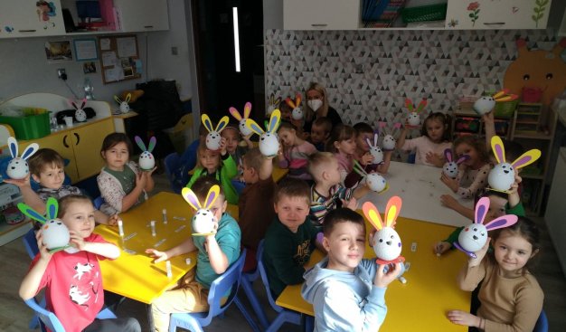 Na zdjęciu grupa dzieci w wieku przedszkolnym, siedzących przy przedszkolnych stoliczkach i trzymający w dłoniach wykonane przez siebie ozdoby wielkanocne w formie królików.