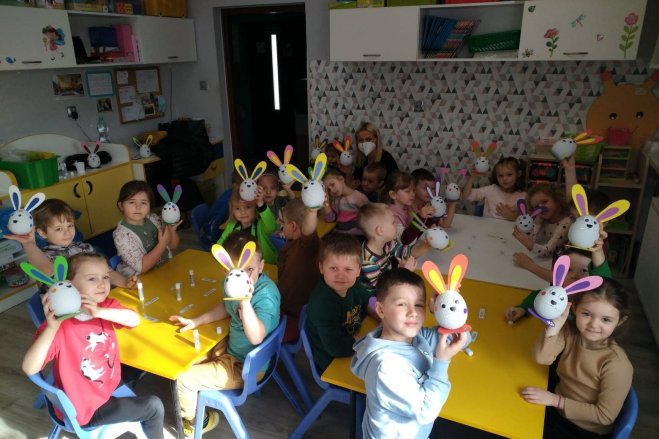 Na zdjęciu grupa dzieci w wieku przedszkolnym, siedzących przy przedszkolnych stoliczkach i trzymający w dłoniach wykonane przez siebie ozdoby wielkanocne w formie królików.