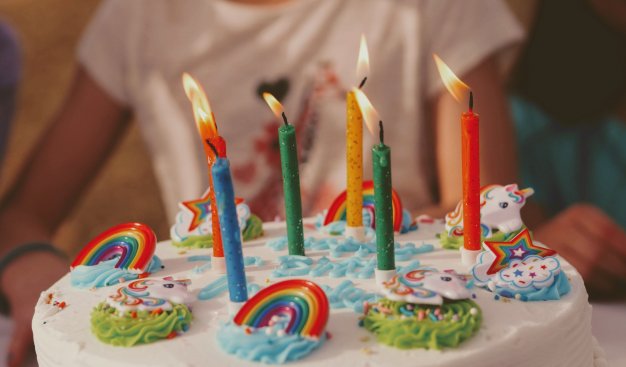 Zbliżenie na tort, udekorowany w stylu dziecięcym - kolorowe tęcze, chmurki i jednorożce,, na środku tortu sześć różnokolorowych świeczek. W tle za tortem widoczna zamazana sylwetka dziecka.