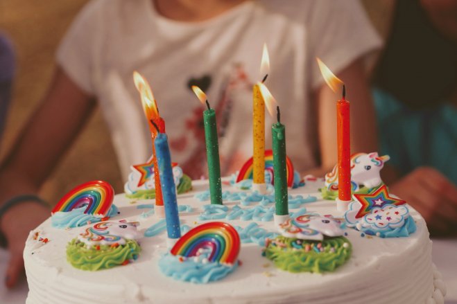 Zbliżenie na tort, udekorowany w stylu dziecięcym - kolorowe tęcze, chmurki i jednorożce,, na środku tortu sześć różnokolorowych świeczek. W tle za tortem widoczna zamazana sylwetka dziecka.