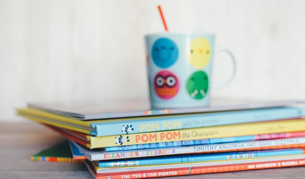 Na stole leży stosik kilkunastu cienkich książek z tytułami w języku nagielskim na grzbietach. NA książkach stroi kolorowy kubek dziecięcy z ikonkami buziek z różnymi minami.
