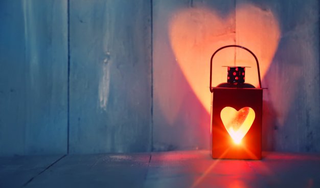 Na tle niebieskich desek stoi lampion z wycięciem w kształcie serca. Płomień świecy wewnątrz  lampionu rzuca z tyłu na ścianę jasny kształt serca.