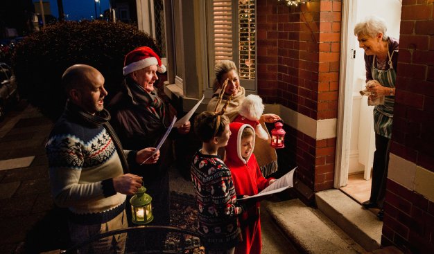 Grupa kolędujących osób stoi pod drzwiami domu. Mają świąteczne przebrania, trzymają w rękach śpiewniki i lampiony.  W progu domu wita ich uśmiechnięta starsza kobieta.