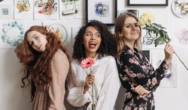 Trzy kobiety w ładnych strojach, trzymają w dłoniach kwiaty i pozują uśmiechnięte do zdjęcia. Za nimi ściana z mnóstwem różnych obrazków w ramkach.