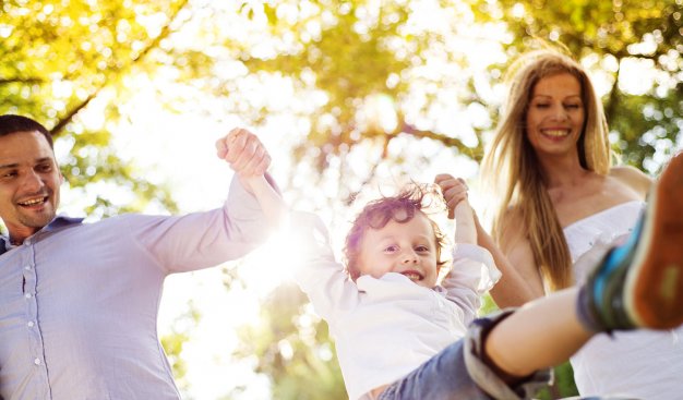 Zdjęcie rodziny. W środku usmiechnięte dziecko, któe trzyma za ręce mężczyznę i kobietę ustawionych po obu jego stronach. Dzień, słoneczna pogoda.