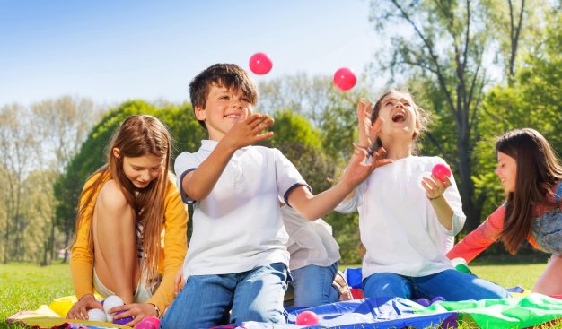 Grupa dzieci w słoneczny dzień bawi się w plenerze podrzucając i łapiąc kolorowe piłeczki.