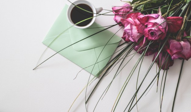 Na jasnym stole leży bukiet kwiatów - róż, filiżanka z kawą oraz list w eleganckiej pastelowej kopercie.