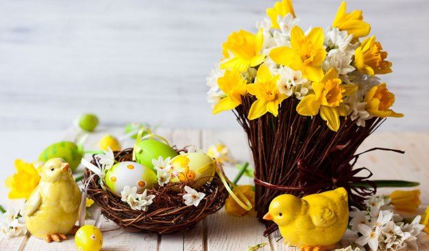 Wielkanocny stroik z żonkili. Obok wiklinowy koszyczek z pisankami. Obok żółte kurki.