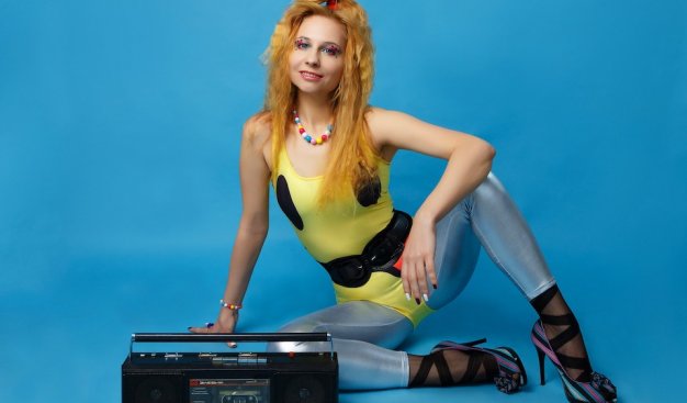 Młoda kobieta w stroju disco z lat 80-tych, z magnetofonem na tle błękitnej ściany.