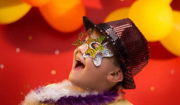 Portret radosnego dziecka przebranego w kolorową maskę i kapelusz z cekinami, na tle czerwonej ściany z balonami. Nastrój urodzinowy.