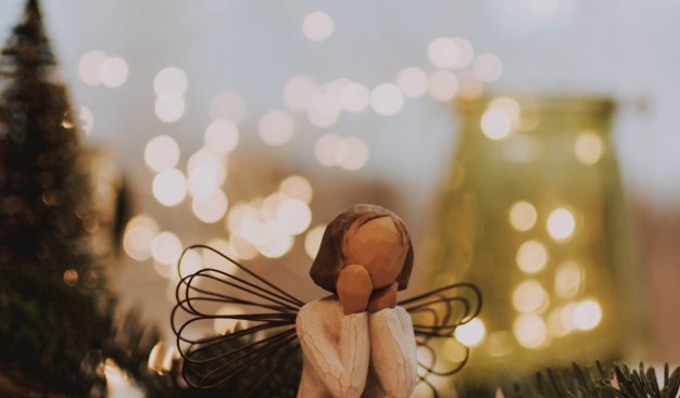 Drewniana figurka aniołka, który siedzi na drewnianym pieńku i podpiera głowę rękoma. NA plecach ma wykonane z drucików skrzydła.  W tle świąteczne światełka i ozdoby - szyszki, choineczki, gwiazdki.