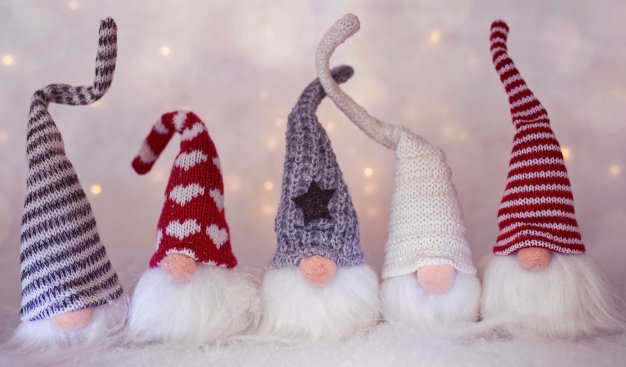 Pięć skrzatów wykonanych z włóczki, ubranych w długie szpiczaste w wełniane czapki w zimowe i świąteczne wzory, umieszczone na jasnym tle błyszczącym świątecznymi światełkami.