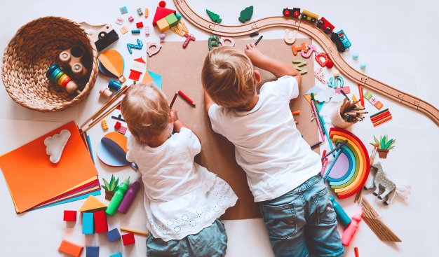 Widok z góry na dwójkę dzieci leżących na podłodze. Wokół nich różnorodne drewniane kolorowe klocki i zabawki, kartki kolorowego papieru i inne akcesoria do zabawy.