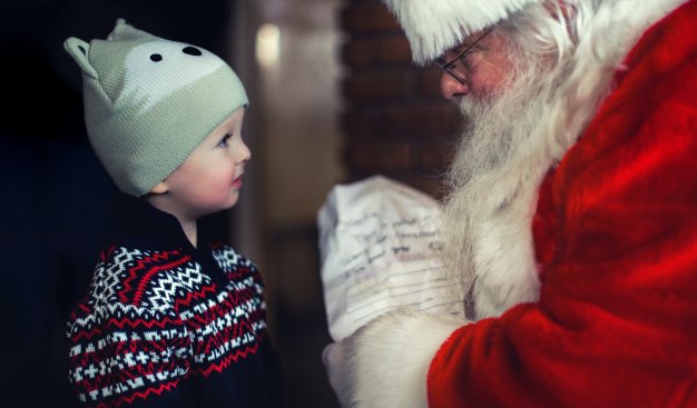 Chłopiec ubrany w ciepły zimowy sweter i czapkę uśmiecha się do ubranego w czerwony strój Mikołaja, który wręcza dziecku paczuszkę,