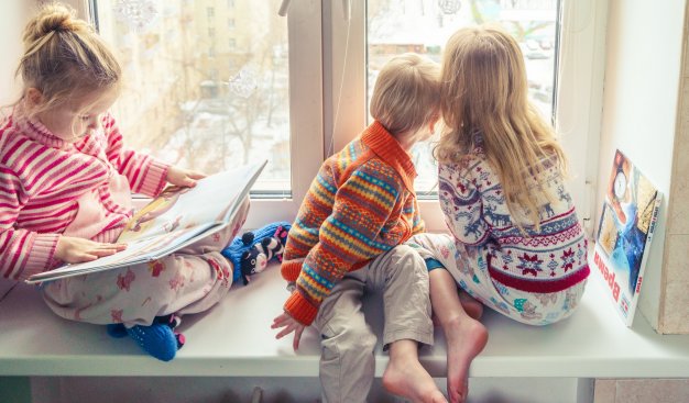 Dzieci ubrane w ciepłe sweterki z motywem zimowym, siedza na szeokim paprapecie okno i spoglądaja za okno na zimowy świat.