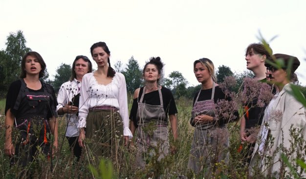Grupa kobiet i mężczyzn stojąca w wysokiej trawie. Ubrana w stroje ludowe.