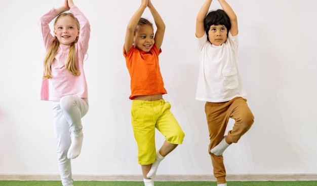 Troje dzieci, dziewczynka i dwóch chłopców, w sportowych strojach, stoi równo w rzędzie wykonując pozycje z jogi.