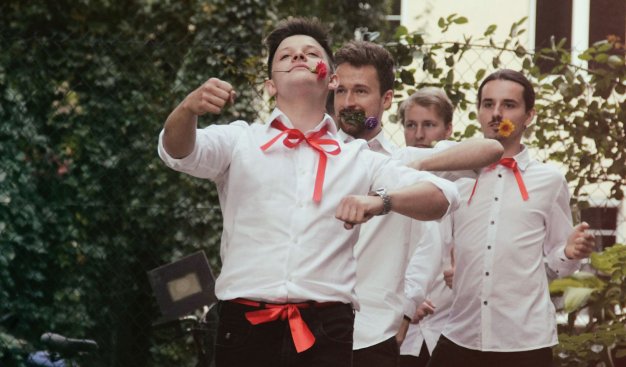 Grupa czterech mężczyzn w białych koszulach i czerwonych wstążkach zawiązanych pod szyją. Idą tanecznym krokiem gęsiego, w zębach trzymając kwiaty