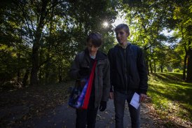 dwóch chłopców w parku, jeden trzyma telefon, baj zerkają w telefon, są skupieni, mają płócienne torby z logiem festiwalu "Kryptonim Lem", wokół zieleń, słońce przebija się przez drzewa