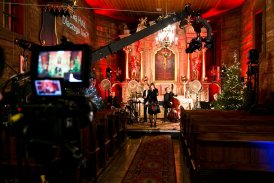Wnętrze małego drewnianego kościoła. Przed ołtarzem śpiewa Anna Branny. Zdjęcie zrobione z oddalenia, widać ramię wysięgnika kamery oraz ekran kamery z rejestrowanym obrazem.