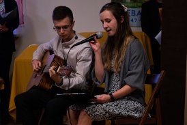 Zdjęcie dwóch osób siedzących, jedna z nich - chłopak gra na gitarze, druga - dziewczyna śpiewa do mikrofonu