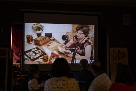 Zdjęcie przedstawiające ekran na którym wyświetlany jest film, na pierwszym planie siedzą ludzie (tyłem do obiektywu).