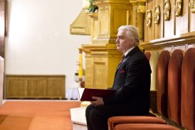Zdjęcie przedstawia faceta, który siedzi w kościele. W rękach trzyma czerwoną teczkę.