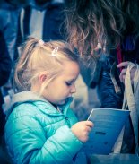 Fotografia przedstawia dziecko w tłumie, które spogląda na książeczkę. Dziewczynka jest zainteresowana książeczką.