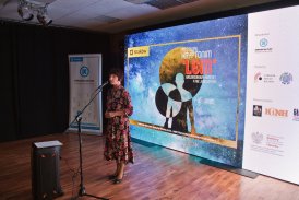 scena, na scenie stoi kobieta, stoi bokiem, mówi do mikrofonu, po prawej stronie na wielkim ekranie wyświetlane jest logo festiwalu "Kryptonim Lem", po lewej stronie ekranu stoi plakat z logiem Ośrodka Kultury Krakow-Nowa Huta