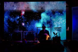 na zdjęciu jest ciemno, jedynym oświetleniem jest ekran, na ekranie wyświetlone jest zdjęcie kosmosu, przed ekranem siedzi mężczyzna, jest skupiony, za nim są różne instrumenty, dzwonki, gongi
