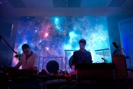 ujęcie z dołu, dwaj mężczyźni siedzą na ziemi przy syntezatorach, są zajęci muzyką, skupieni, z tyłu wyświetla się zdjęcie kosmosu, światło w pomieszczeniu jest niebieskie