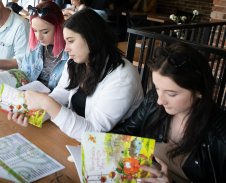Zbliżenie na siedzące przy drewnianym stole trzy uczestniczki zajęć - młode dziewczyny przeglądające książki dla dzieci pt. "Nowohuckie podróże Pana Dyni".
