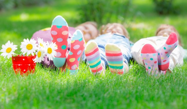 Kolorowe dziecięce skarpety w paski i groszki. Dzieci leżą na zielonej trawie. Obok nich czerwony dzbanuszek z letnimi kwiatami.