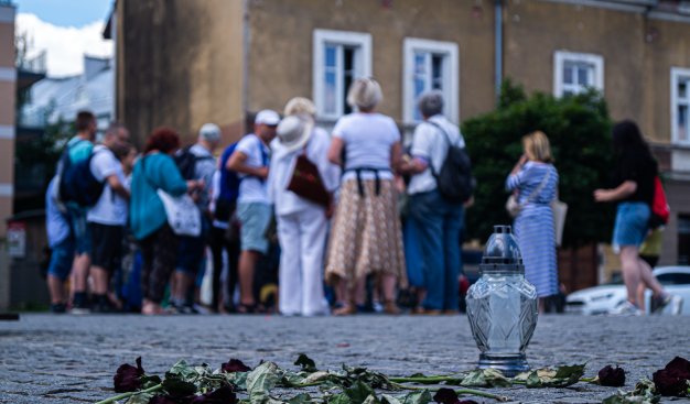 Zdjęcie przedstawiające grupę ludzi stojących tyłem, na pierwszym planie znajduje się znicz stojący na bruku oraz uschnięty kwiat