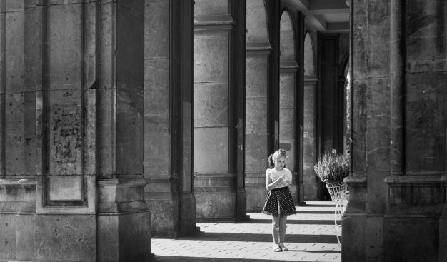 Czarno biała fotografia. Słoneczny dzień, pod arkadami w pobliżu Placu Centralnego stoi dziewczynka z warkoczykami i tiulowej spódniczce w grochy. Fotografia Marcina Koleśnika.