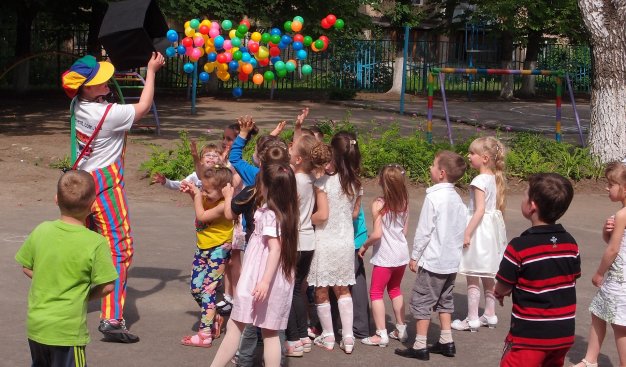 Słoneczny dzień. Animator bawiący się z grupą dzieci. Z trzymanego nad głowami dzieci ciemnego pudła wyrzuca małe kolorowe kulki, które dzieci próbują łapać. Widać ruch.