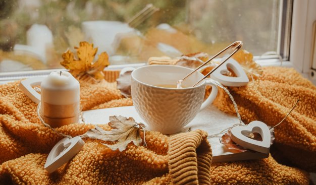 Jesienna kompozycja ułożona na parapecie okna. Na grubym rudym swetrze stoją filiżanka z parzaca się herbatą na podstawce, jesienne liście, świeczka oraz światełka w kształcie serduszek wykonanych z drewna.
