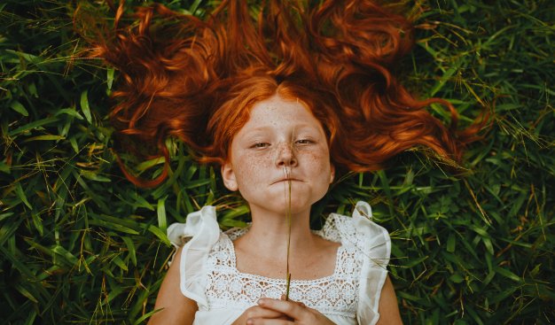 Dziewczynka z długimi rudymi włosami i piegami na twarzy, ubrana w białą bluzkę z falbankami leży na zielonej trawie. W dłoniach trzyma źdźbło trawy, które przygryza wargami.