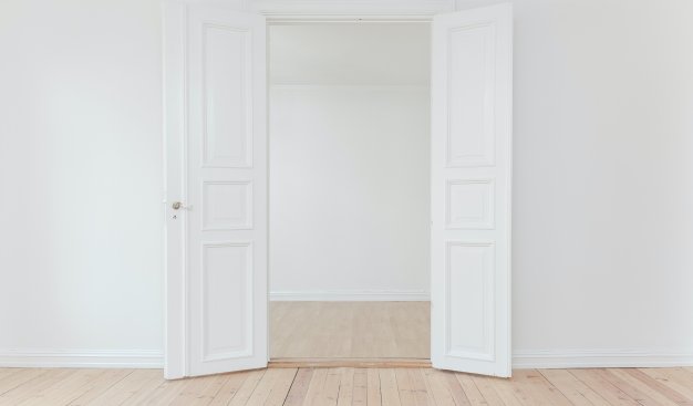 Sala z białymi ścianami i drewnianą podłogą. Centralnie szeroko otwarte dwuskrzydłowe białe drzwi. Za nimi widać drugą białą ścianę i fragment drewnianej podłogi.