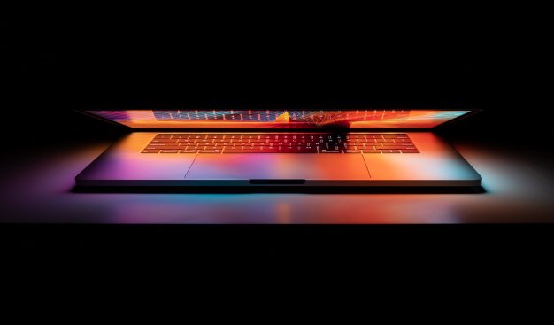 Częściowo przymknięty laptop leży na stole. Feeria kolorów z wyświetlacza odbija się w klawiaturze i dookoła komputera. W tle ciemność.