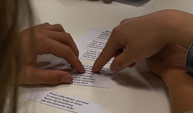 Zdjęcie pzredstawiająjce dłonie układające kartki na stole - kartki zawierają tekst wierszy, które należy ułożyć w całość