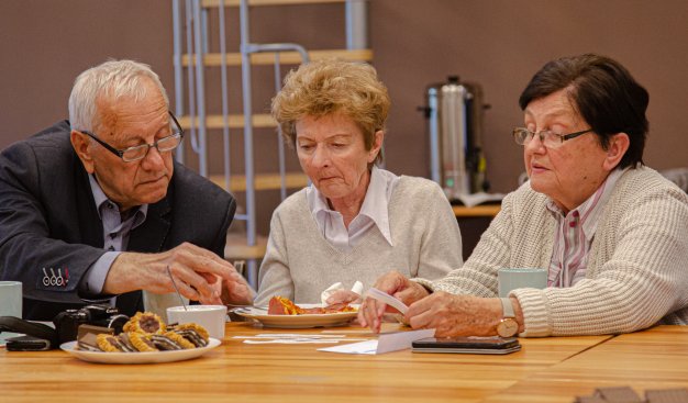 Zdjęcie przedstawiające trzy osoby siedziące przy stole: starszego mężczyznę w okularach, ubranego w ciemną marynarkę, starszą kobietę w białej koszuli z krótkimi włosami, starszą kobieteę w okularach z cienymi włosami. Wszytskie osoby czytają tekst, sied