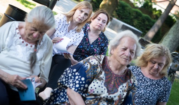 Zdjęcie wykonane podczas warsztatów plenerowych. Na pierwszym planie siedzi pięć kobiet w różnym wieku, każda z leksykonem w ręku. Kobiety zasłuchane. W tle pnie drzew i widoczna mała infrastruktura parku - ławki, murki.