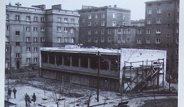 Czarno-białe zdjęcie, w tle bloki mieszkalne, na pierwszym planie jednopiętrowy budynek Klubu Jędruś w stanie surowym.