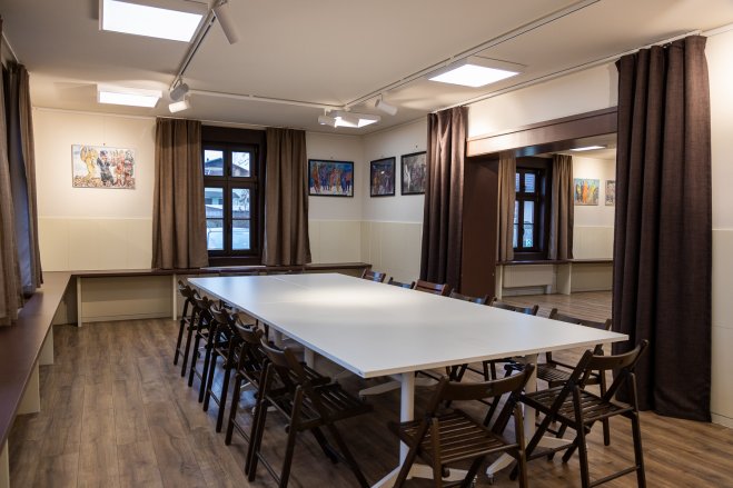 Zdjęcie przedstawiające jasne wnętrze Klubu Krzesławice. Na środku pomieszczenia duzy stół z klkudziesięcioma krzesłami wokół.
