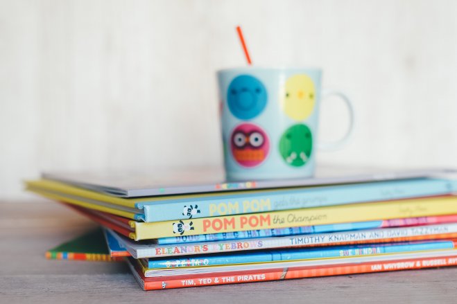 Na stole leży stosik kilkunastu cienkich książek z tytułami w języku nagielskim na grzbietach. NA książkach stroi kolorowy kubek dziecięcy z ikonkami buziek z różnymi minami.