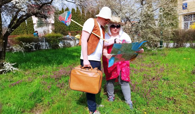 Dwie osoby stojące na trawie wśród drzew. Obie ubrane jak turyści, z kapeluszami na głowach, z walizką w ręku i trzymaną w rękach rozłożoną mapą.