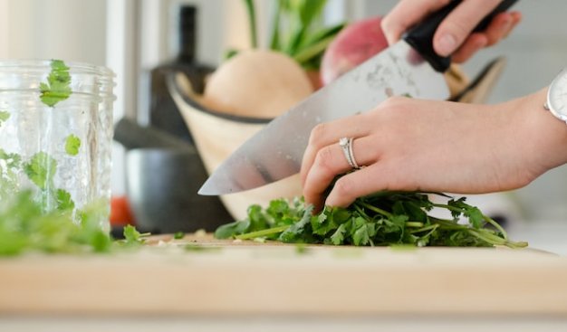 Zdjęcie przedstwiające dłonie kobiety krojące na desce zioła, w tle znajdują się inne produkty kuchenne