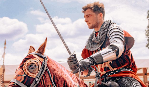 Zdjęcie przedstawiające rycerza w zbroi jadącego na koniu i trzymającego uniesiony miecz.